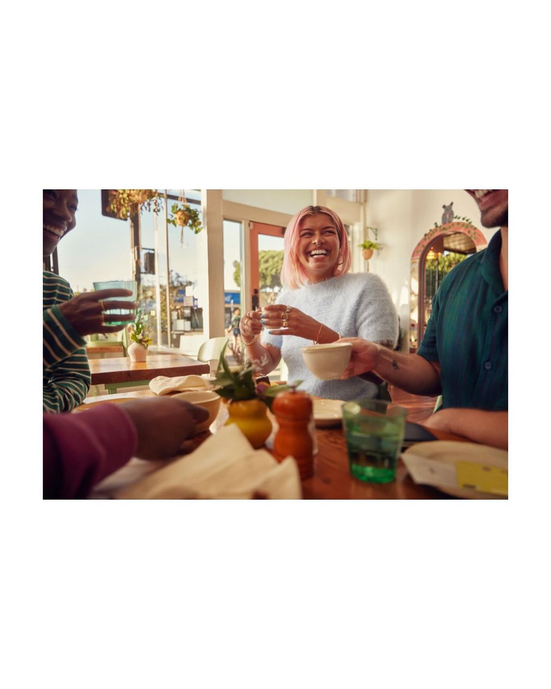 A group of four young adults are sitting at a table with plates and glasses. One person has bright pink hair and is laughing.