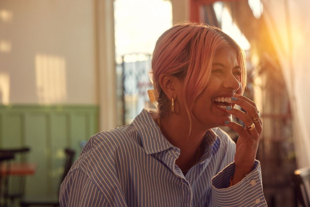 Close-up of a laughing woman with pink hair wearing a blue striped shirt, gold hoop earrings, and multiple rings indoors in a bright, airy setting.