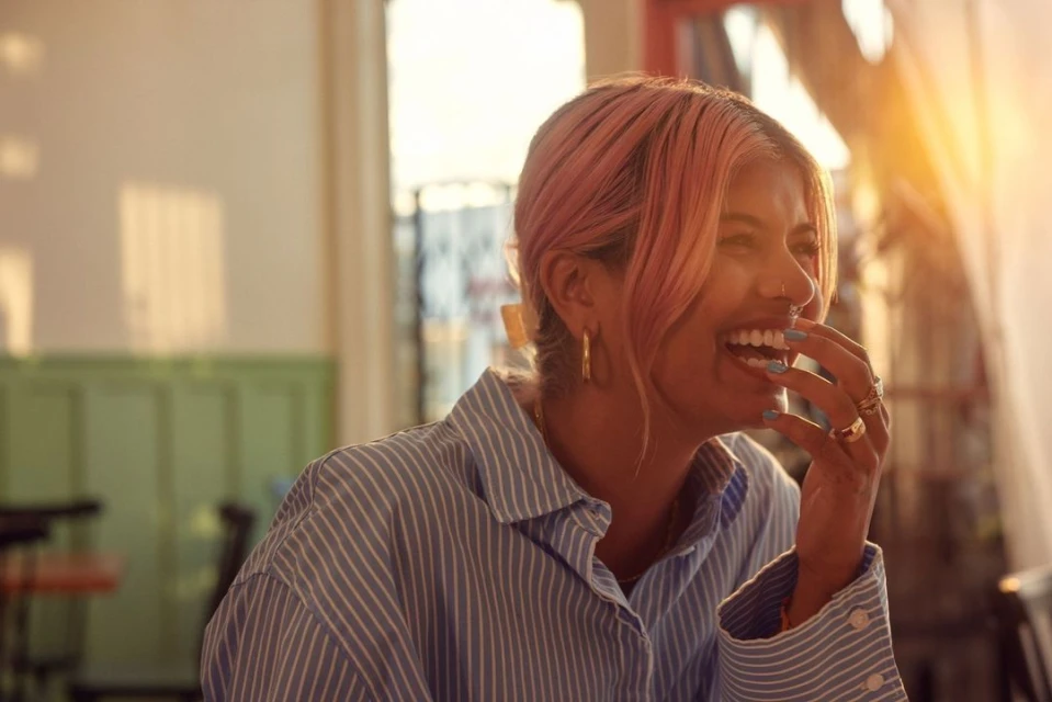 Close-up of a laughing woman with pink hair wearing a blue striped shirt, gold hoop earrings, and multiple rings indoors in a bright, airy setting.