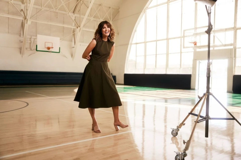 Adult woman in dress smiling and posing in a gymnasium next to lighting equipment with a basketball hoop and large windows in the background.