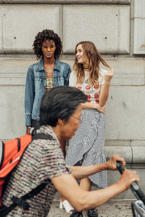 Two young women in stylish summer outfits pose against a concrete wall, with a blurred cyclist in the foreground on an urban street.