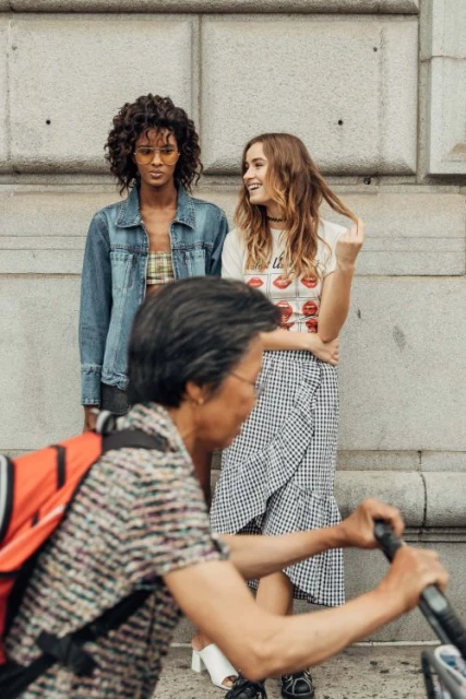 Two young women in stylish summer outfits pose against a concrete wall, with a blurred cyclist in the foreground on an urban street.