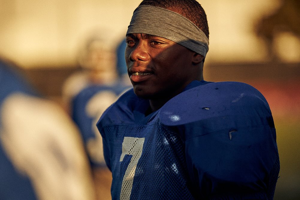 Close-up of an African-American football player in a blue jersey with a gray headband on an outdoor field on a sunny day.