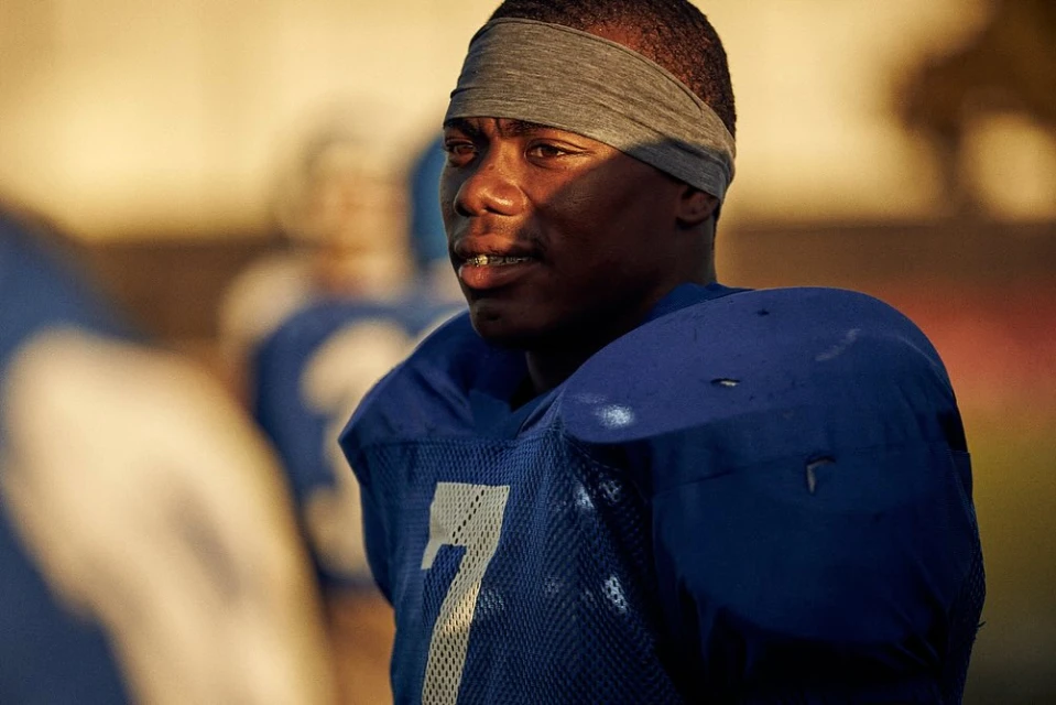 Close-up of an African-American football player in a blue jersey with a gray headband on an outdoor field on a sunny day.