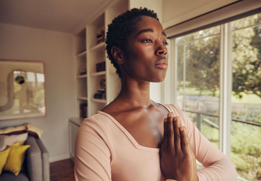 Woman with hands in prayer pose indoors, natural light, wearing pink shirt, window and bookcase in the background, wellness lifestyle.