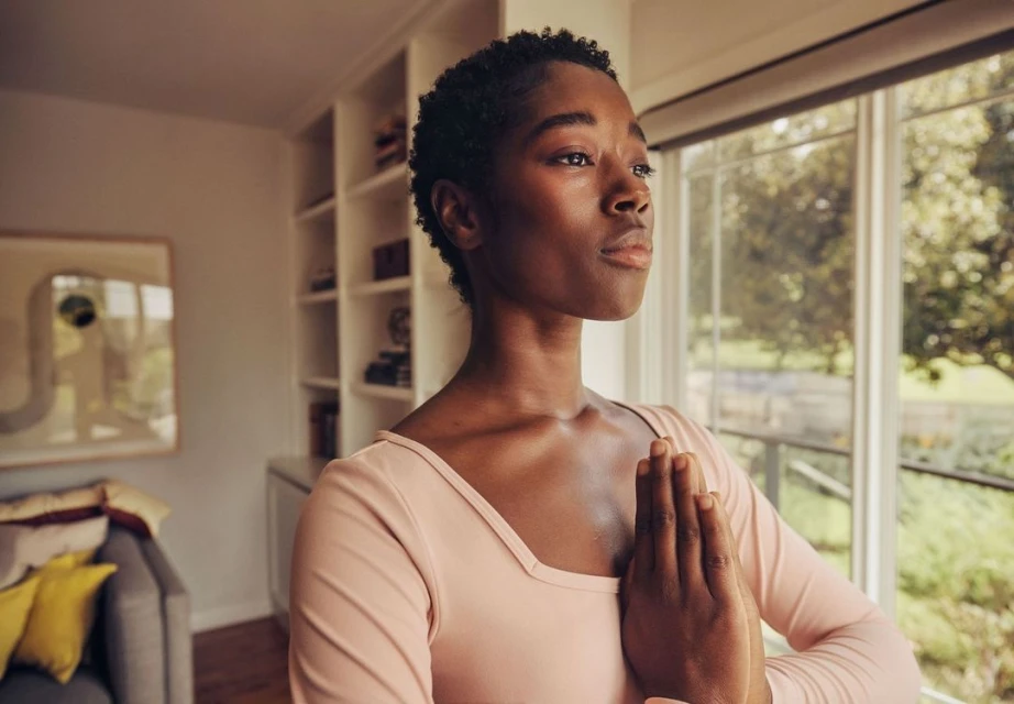 Woman with hands in prayer pose indoors, natural light, wearing pink shirt, window and bookcase in the background, wellness lifestyle.