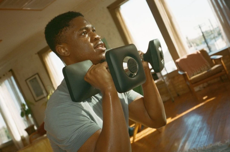 An African American man works out indoors with a grey dumbbell. He is wearing a grey t-shirt and is looking up and to the right.