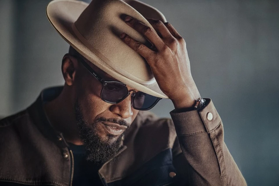 Stylish adult man wearing sunglasses and a fedora hat, captured in a close-up portrait with a blurred grey background.
