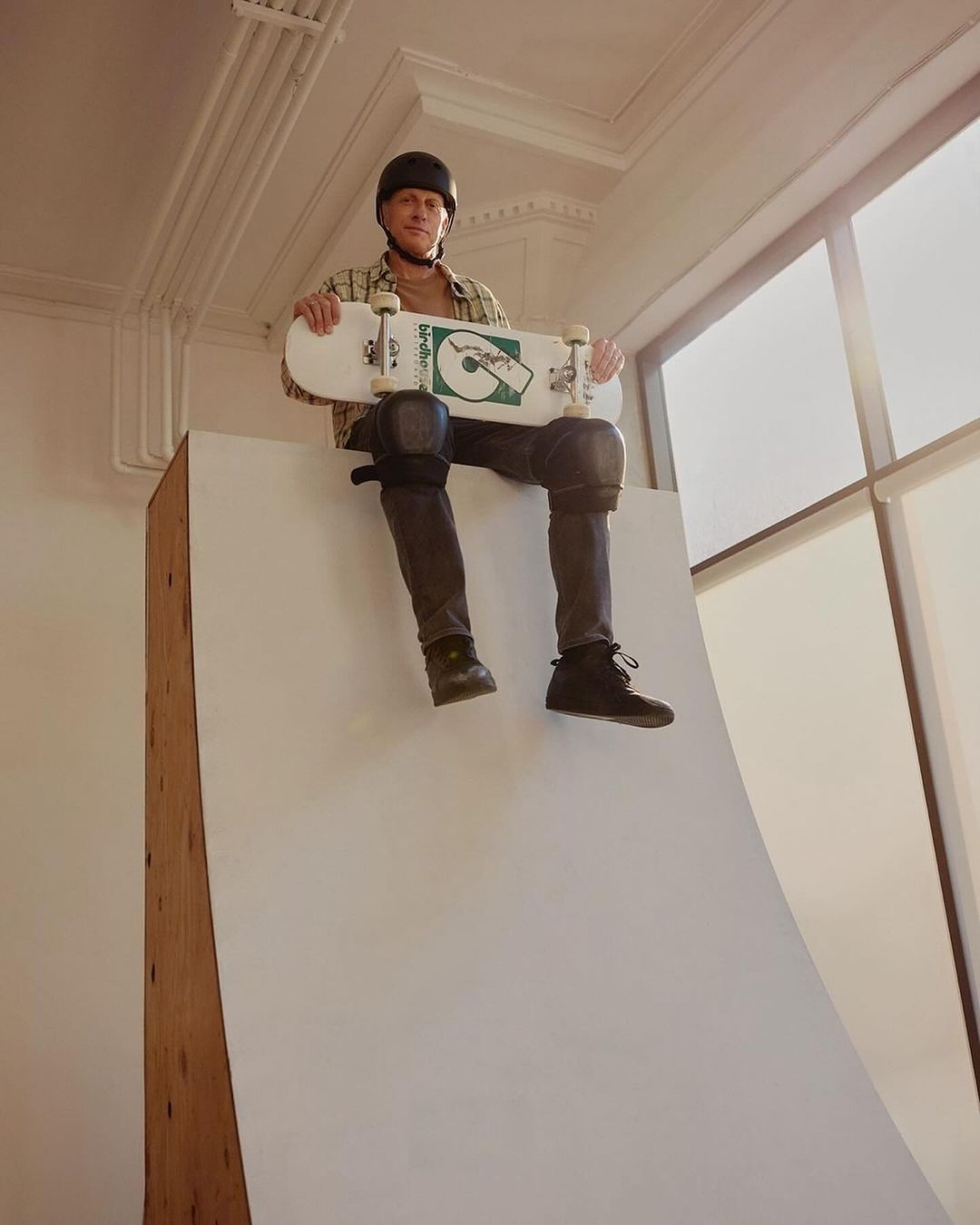 Skateboarder Tony Hawk sits on a white ramp wearing a helmet and knee pads, holding a skateboard in an indoor, well-lit area.