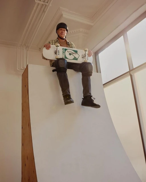Skateboarder Tony Hawk sits on a white ramp wearing a helmet and knee pads, holding a skateboard in an indoor, well-lit area.