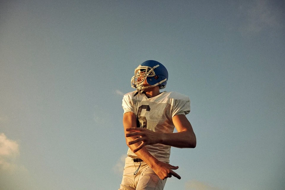 Low-angle shot of a football player in uniform with helmet on, looking up against a pale blue sky with soft clouds, natural light.