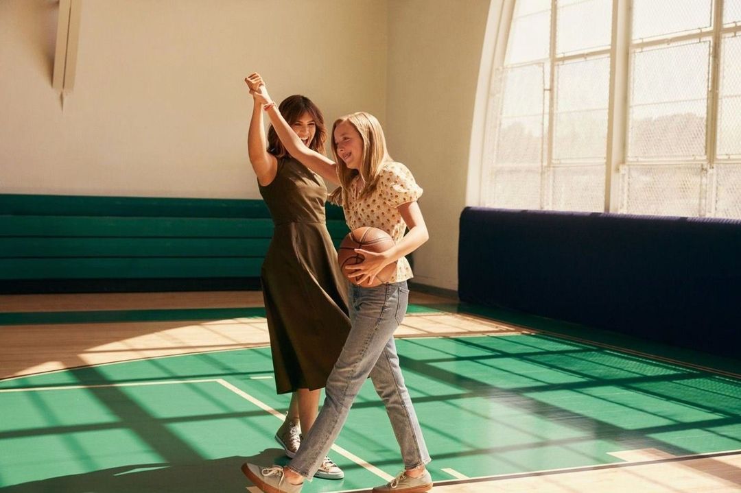 Two women celebrate in a gymnasium, one holding a basketball. The pair are smiling, with shadows cast across the green floor.
