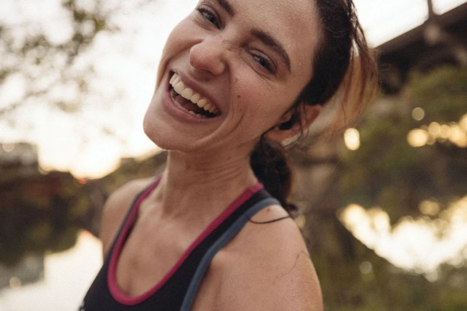 Close-up of a happy adult woman in athletic wear laughing outdoors with blurred greenery in the background and warm lighting.