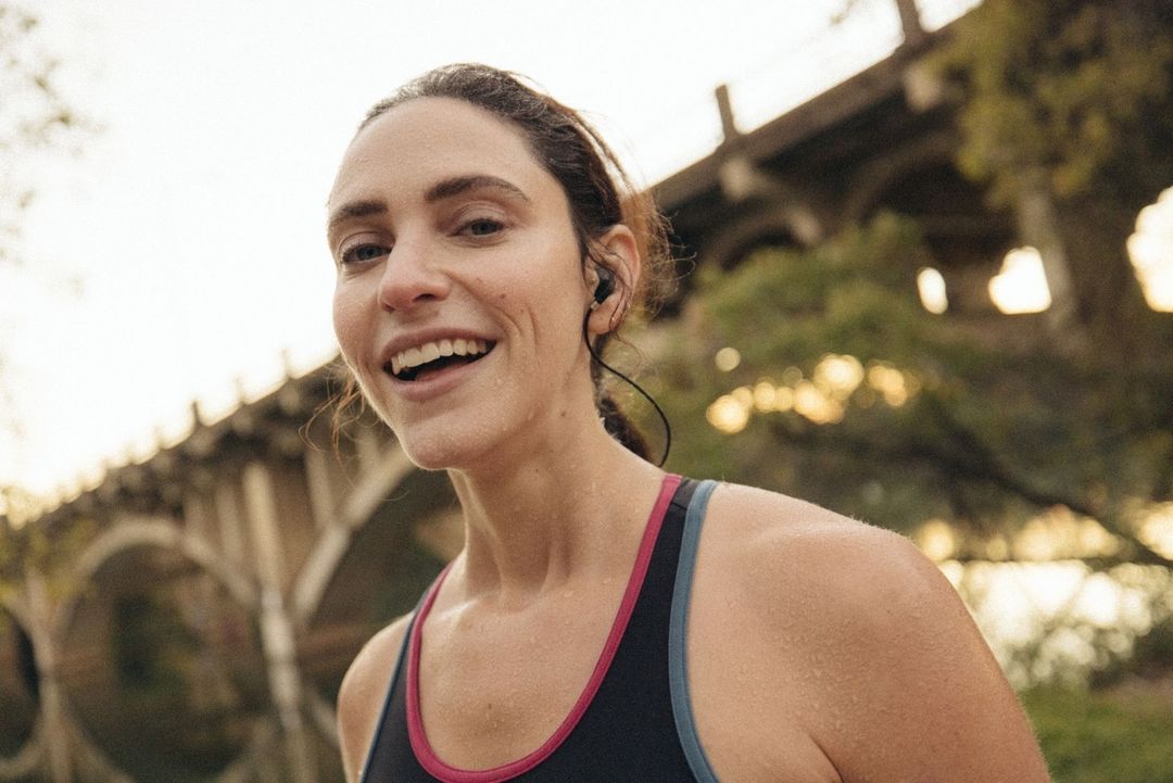 Close-up of smiling woman with earbuds in sports bra under bridge with greenery. Fitness, active lifestyle, healthy living concept.
