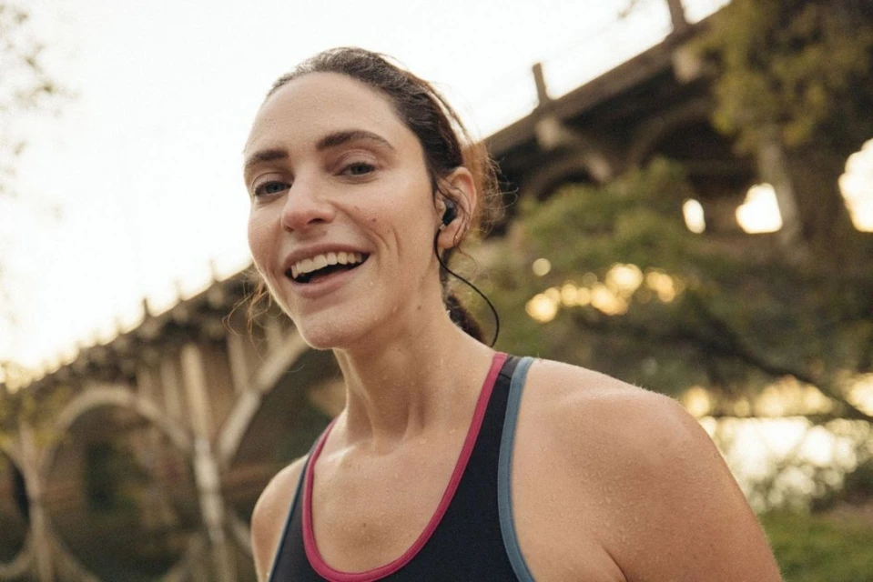 Close-up of smiling woman with earbuds in sports bra under bridge with greenery. Fitness, active lifestyle, healthy living concept.
