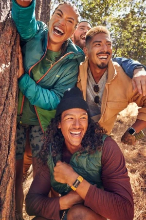 Four diverse adults laugh while standing close together outdoors by a tree; shallow depth of field, natural lighting.