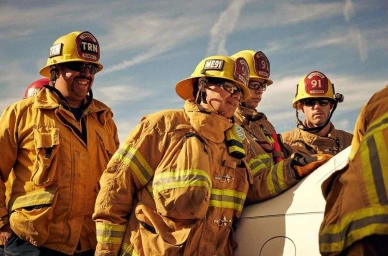 Group of firefighters in yellow turnout coats and yellow helmets smiling outdoors against a clear sky.