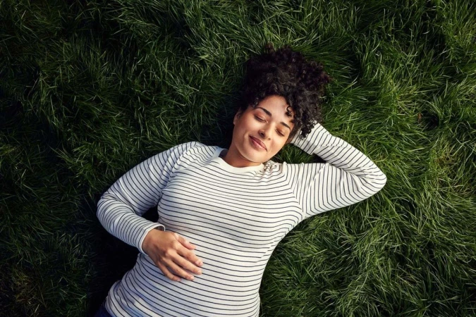 A woman with curly dark hair wearing a striped sweater lying comfortably on green grass with her eyes closed.