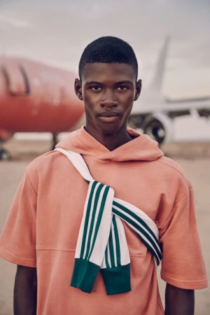 Black man wearing a peach t-shirt and white striped sweater tied across his chest outdoors with an airplane in the background