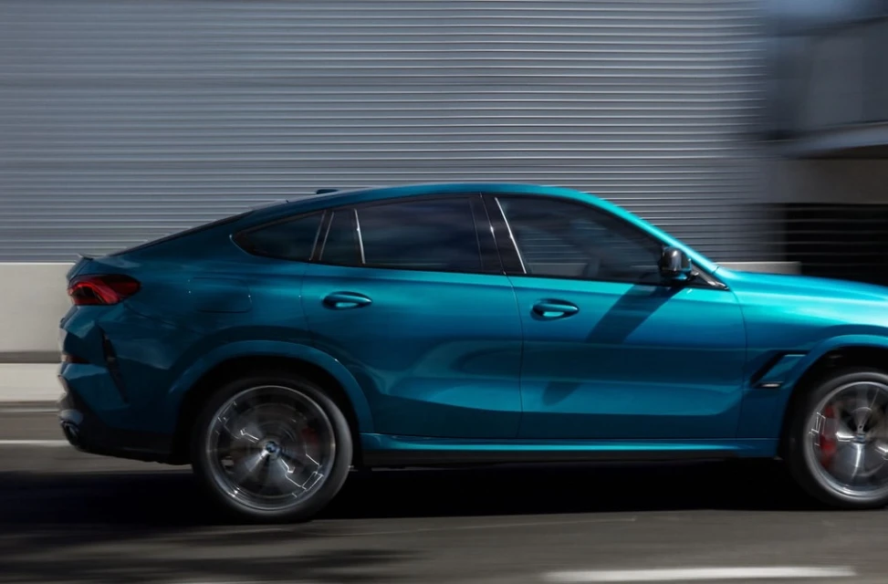 A teal BMW X6 M Competition SUV is captured in motion against a modern building, highlighting its sleek design and dynamic performance.