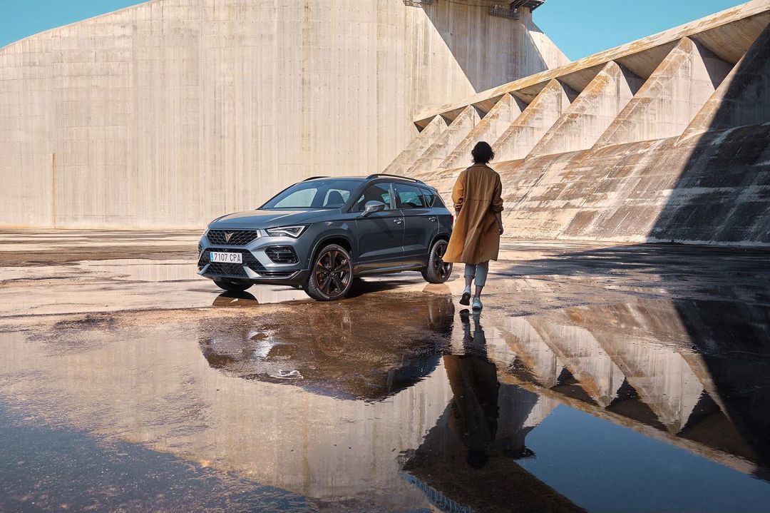 Gray Cupra Formentor SUV parked on a wet surface near a concrete dam, with an adult female figure walking toward the structure.