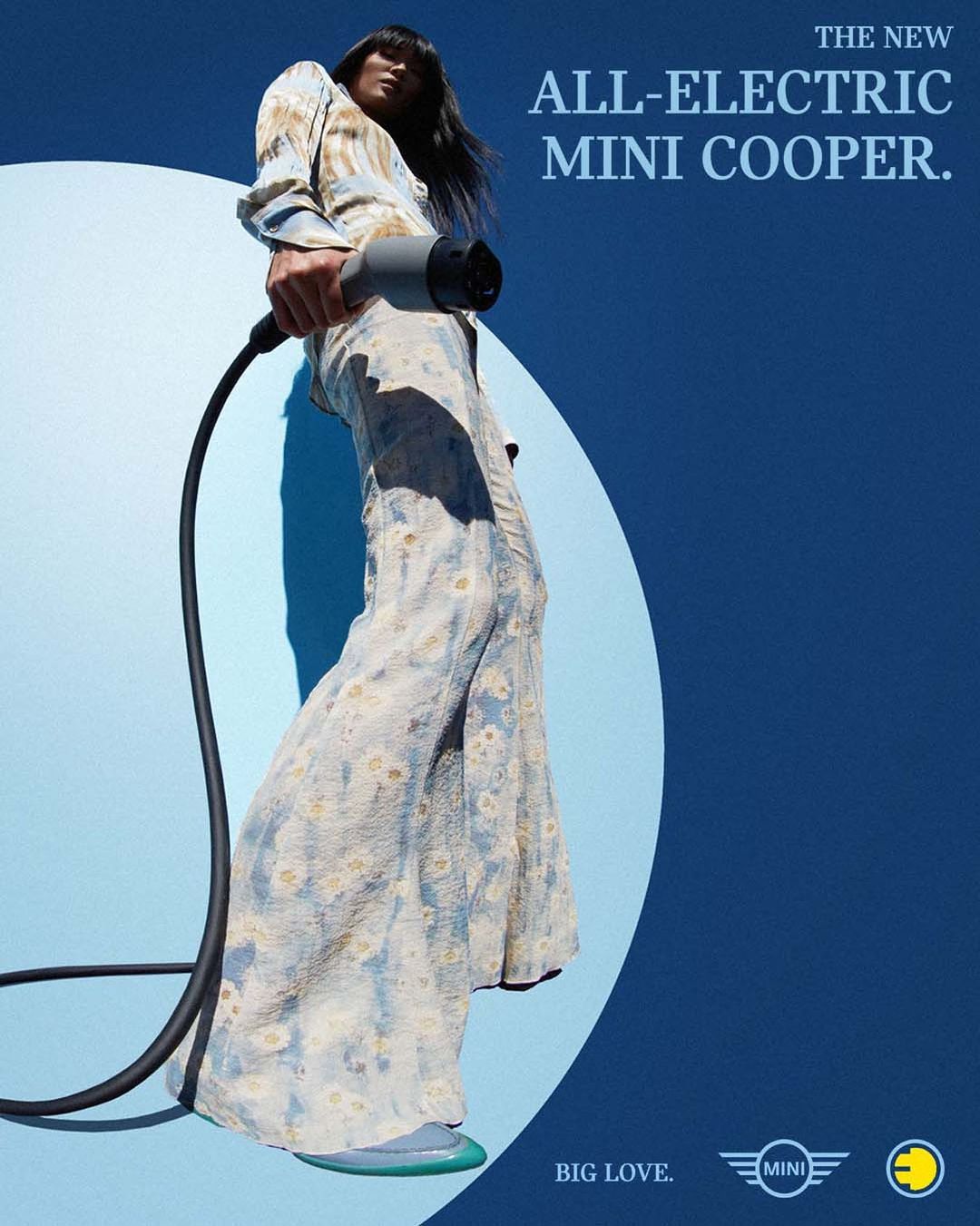 An adult woman in a floral patterned jumpsuit holds a charger for an electric Mini Cooper car against a gradiated blue background.