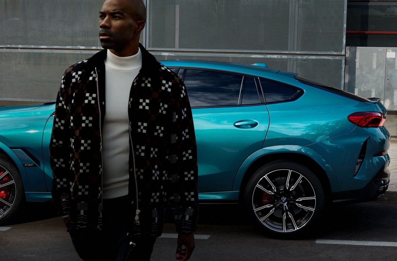 Man in a patterned jacket stands next to a teal BMW X6 on a city street, showcasing a modern urban automotive and fashion scene.