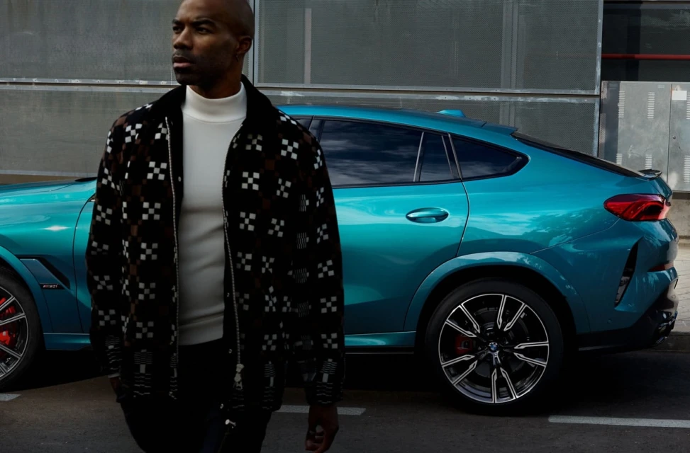 Man in a patterned jacket stands next to a teal BMW X6 on a city street, showcasing a modern urban automotive and fashion scene.