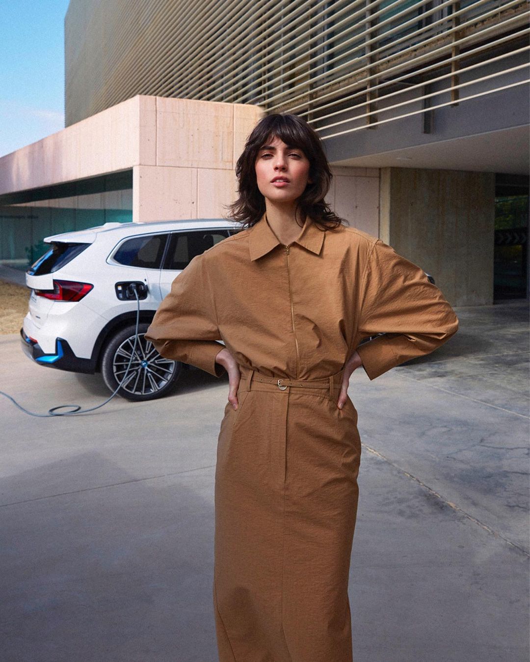 A woman in a tan dress stands in front of a white BMW SUV that is plugged into a charging station against a modern building.