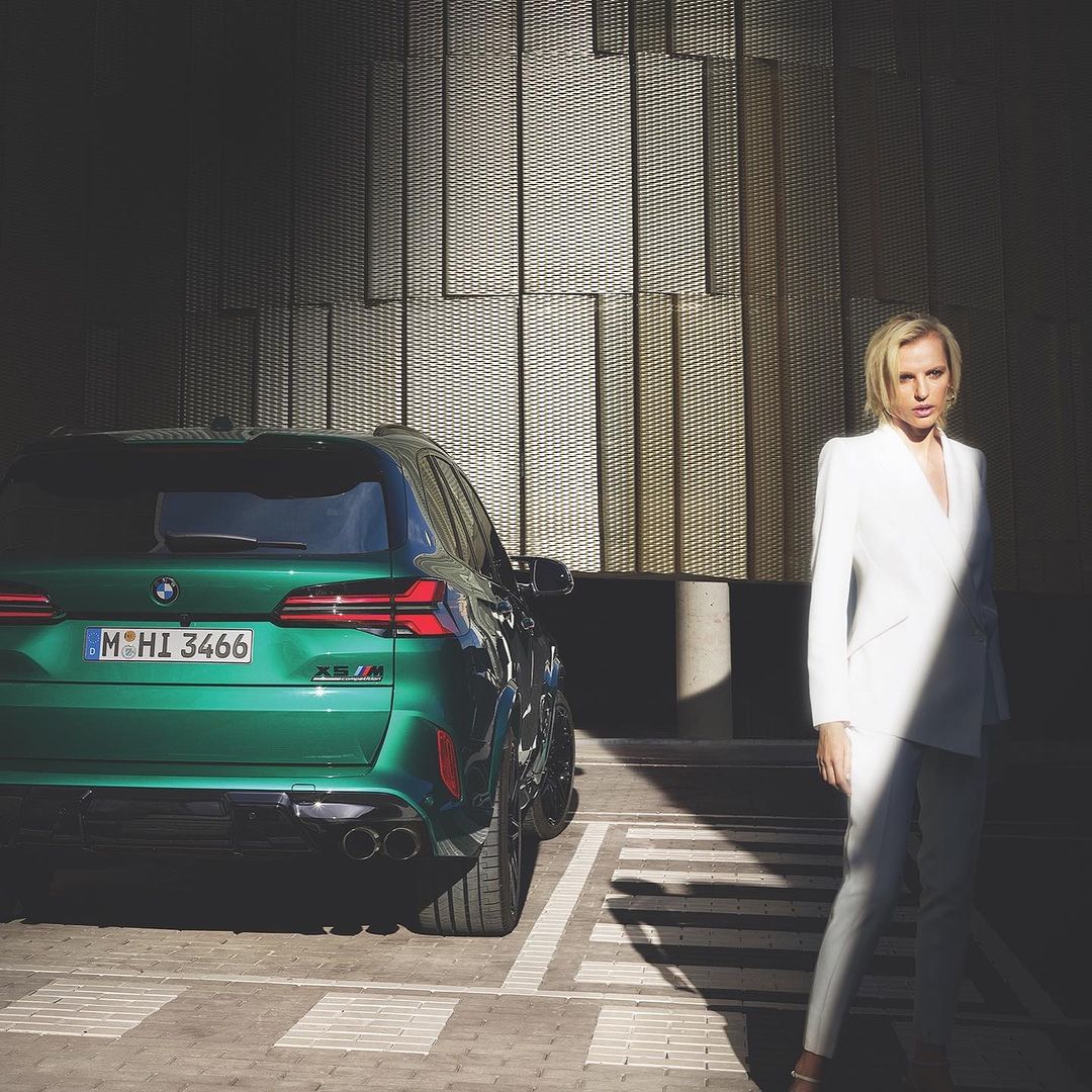 Woman in white suit poses next to a green BMW X5 M parked in a brightly lit parking lot with textured building behind.