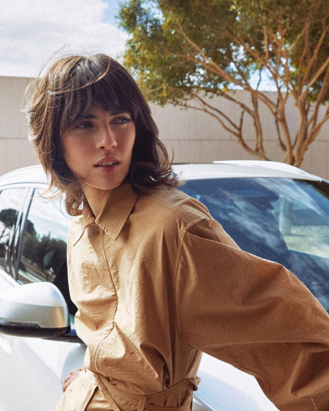 Adult female model poses in tan collared shirt leaning on the front of a white car with green foliage in the background.