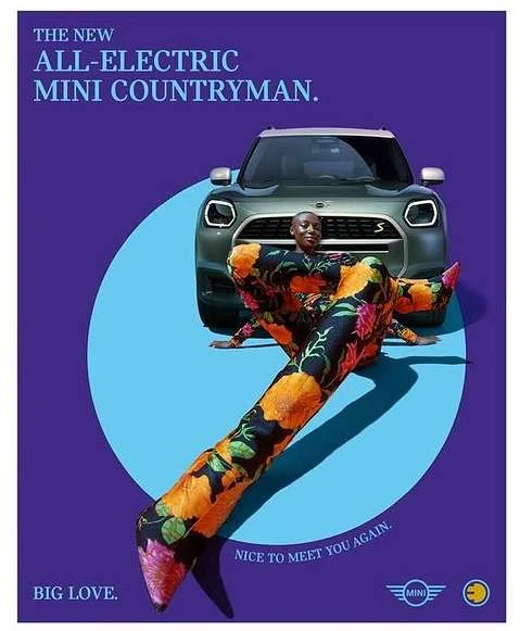 Green Mini Countryman electric car with a black woman in a floral pantsuit posing in front on a light blue and purple background.