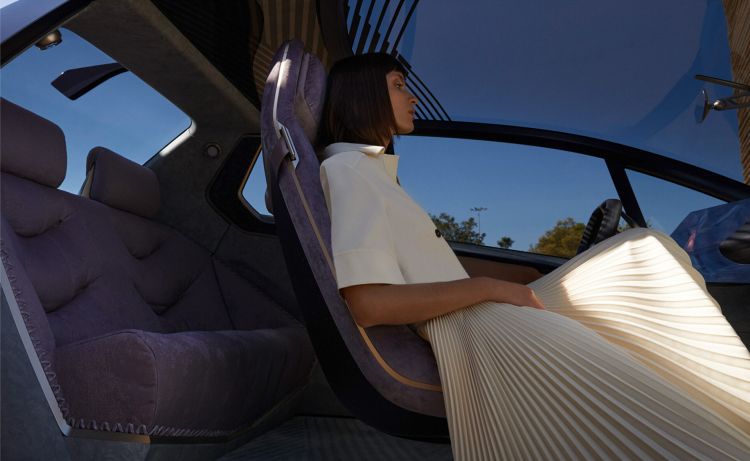 Side view of an adult woman seated inside of a modern car interior with plush lavender upholstery and beige-colored pleated skirt and blouse.