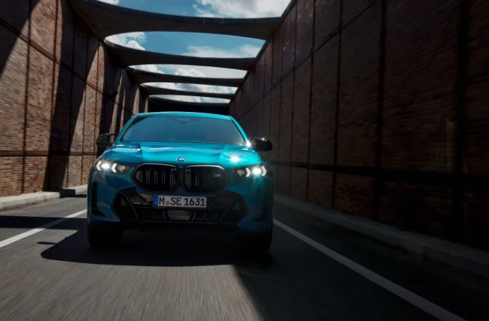 A teal BMW SUV drives toward the camera through a covered tunnel, showcasing its design and presence on the road with brick walls.
