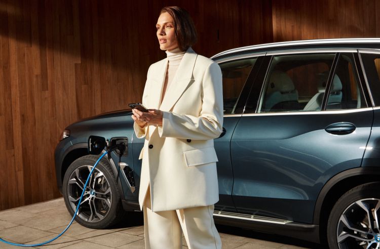 A woman in a white suit stands near a teal BMW electric car plugged in to charge, parked in front of a wood-paneled wall.