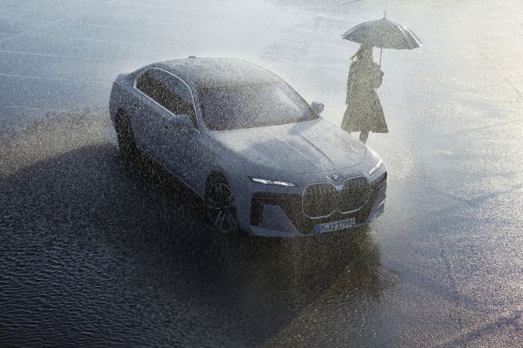Grey BMW car drives on wet asphalt road in the rain, with a person holding an umbrella visible in the background on a bright day.