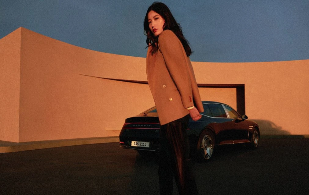 Woman in a tan blazer poses in front of a dark-colored Genesis car with a modern building and soft blue sky in the background.
