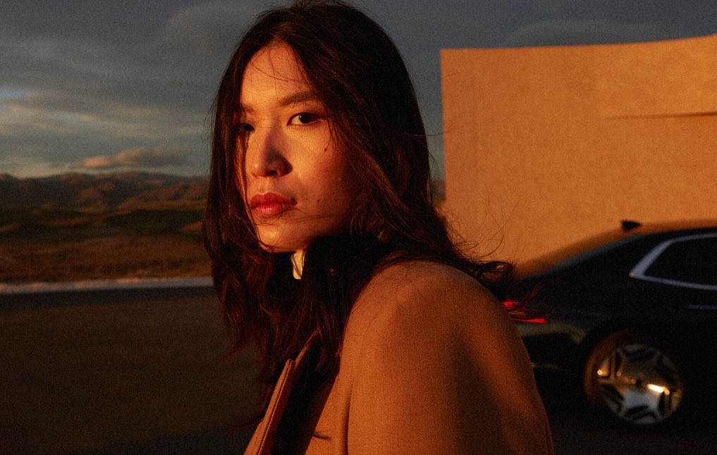 Portrait of an Asian woman in golden hour sunlight outdoors, with a car and building wall in the background.