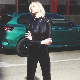 Blonde woman wearing a black sequin jumpsuit posing with arms crossed in front of a green car outdoors.