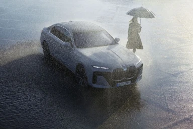 High-angle view of a grey electric car parked in heavy rain while a person holds an umbrella nearby.