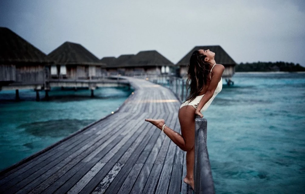 Woman in a swimsuit poses on a wooden boardwalk next to ocean villas. Moody shot of travel and tourism in a tropical resort location.