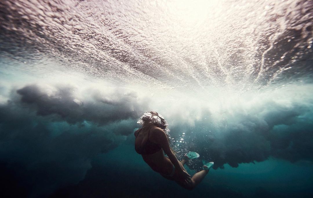 Underwater shot of a woman swimming in the ocean, wearing fins, as the waves crash above her creating dynamic patterns and light.
