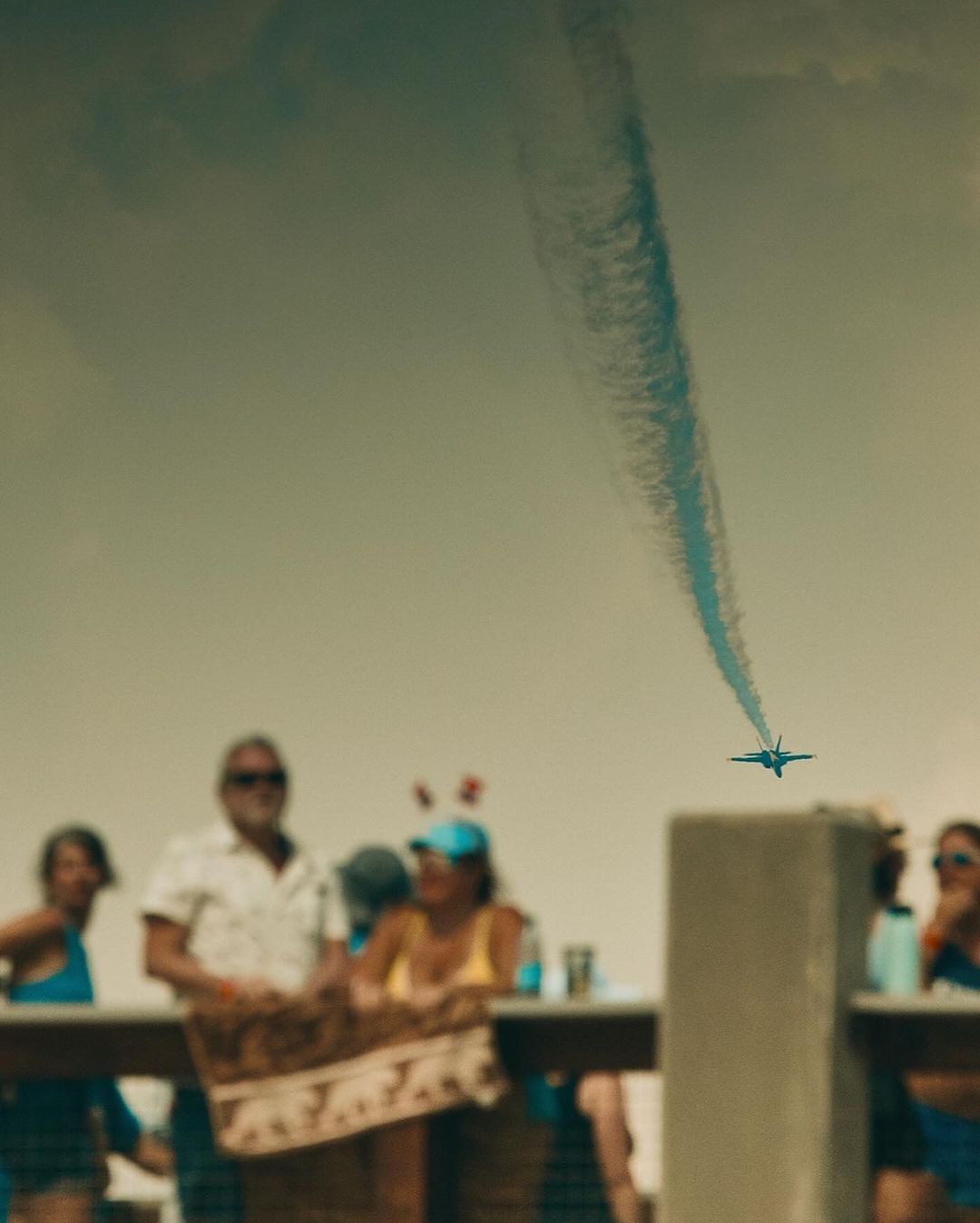 Aerobatic airplane flies over a crowd, leaving a blue contrail against the sky, watched by blurred spectators at an outdoor event.
