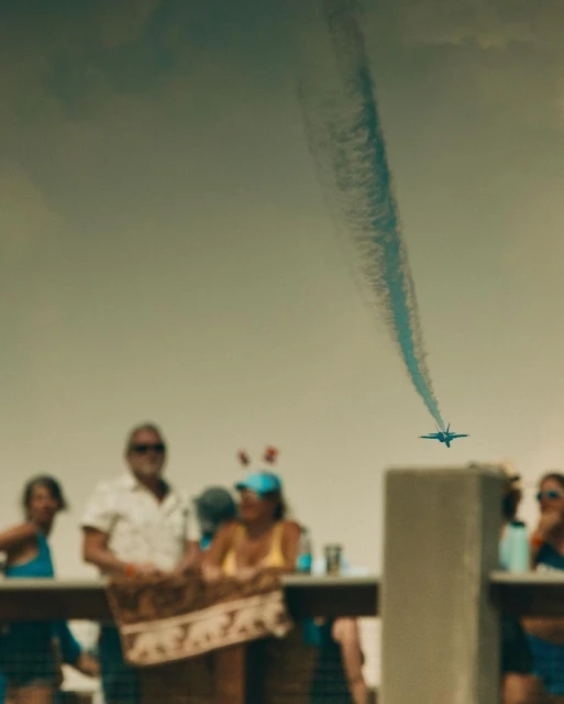 Aerobatic airplane flies over a crowd, leaving a blue contrail against the sky, watched by blurred spectators at an outdoor event.