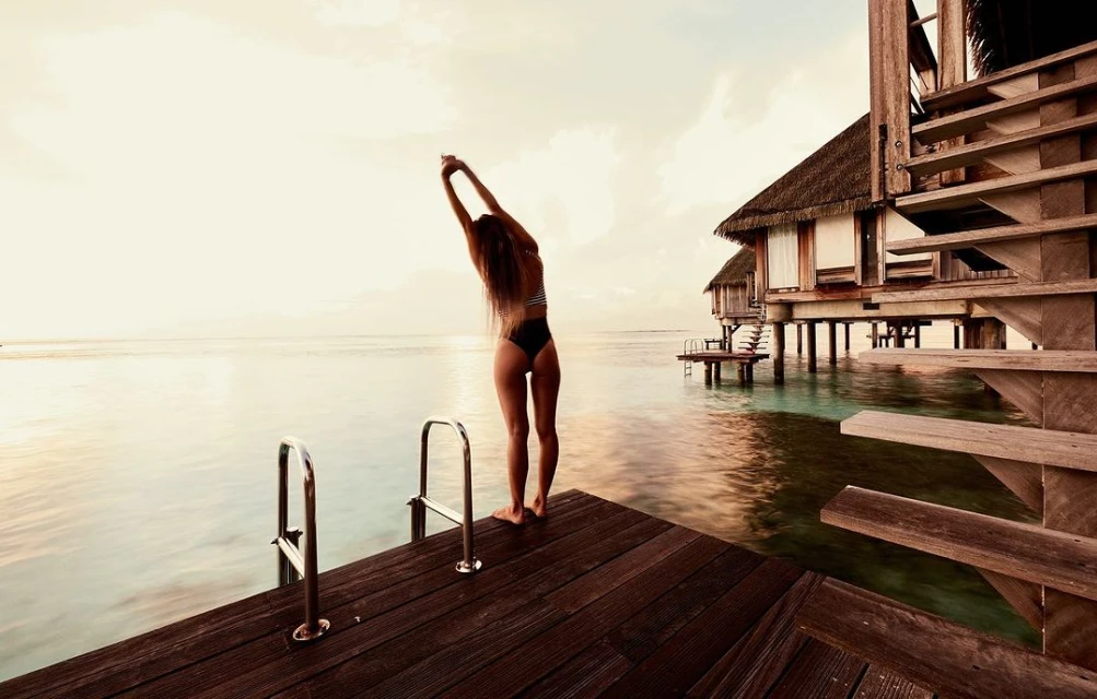 Woman in swimwear stretches on a wooden dock at sunset, near a thatched-roof overwater bungalow. Vacation vibes with ocean views.