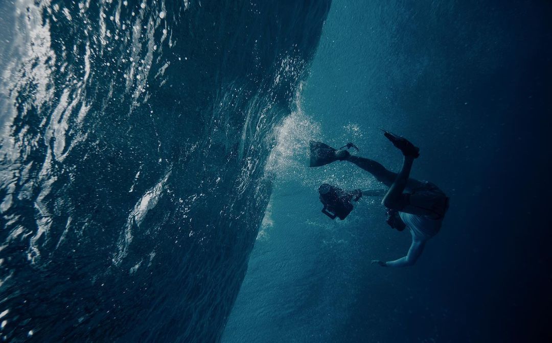 Underwater shot of two divers exploring ocean depths, one using a camera, with sunlight piercing through water's surface creating a serene scene.