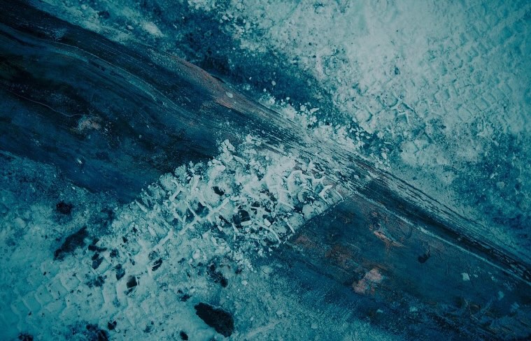Aerial view of textured icy terrain with dark tire tracks running diagonally across frosty ground and snowy patches