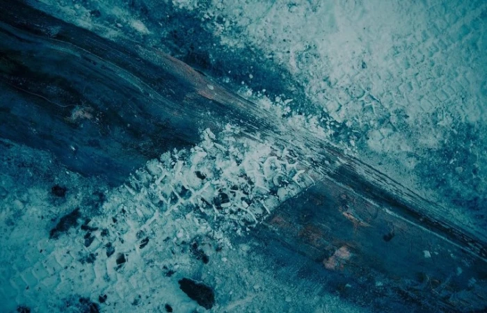 Aerial view of textured icy terrain with dark tire tracks running diagonally across frosty ground and snowy patches