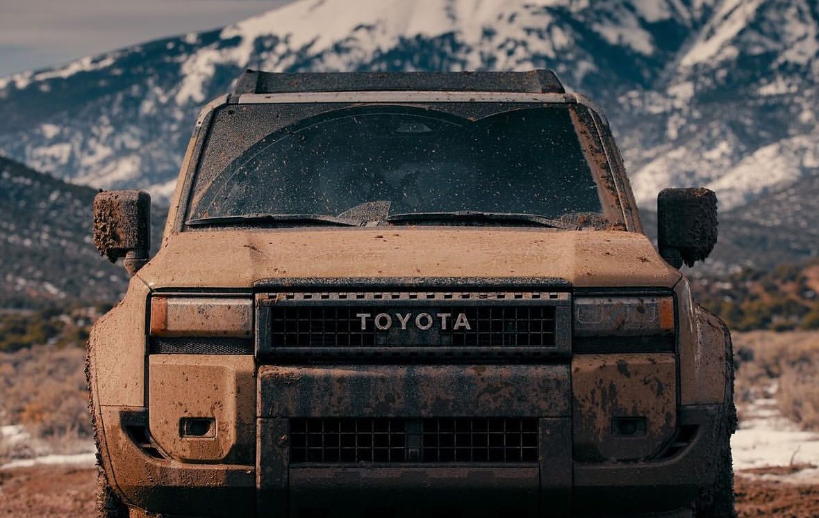 Front view of a tan Toyota SUV covered in mud, parked on a dirt road with snow-capped mountains in the background on a cloudy day.