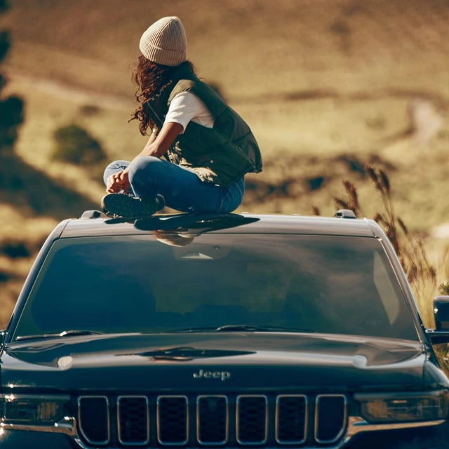 Woman sits atop a black Jeep Grand Cherokee in a rural landscape under warm lighting, showcasing the car's design and outdoor lifestyle.